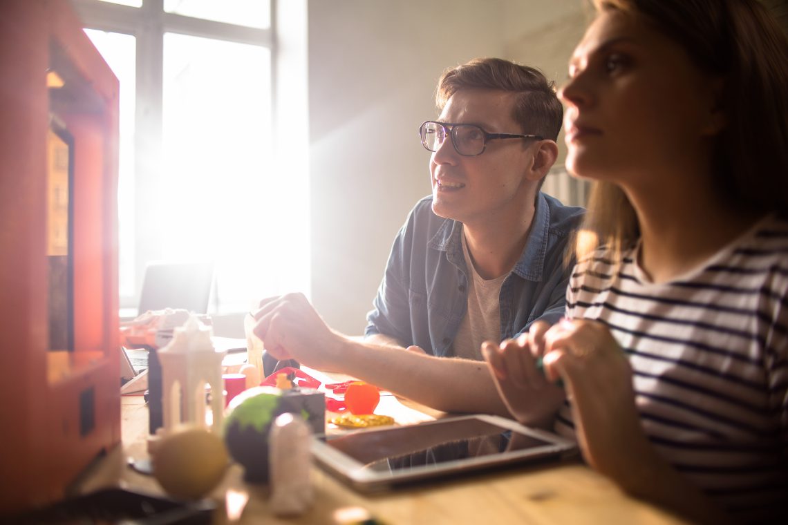 Curious university students sitting at wooden desk and keeping eye out for process of 3D printing, lens flare