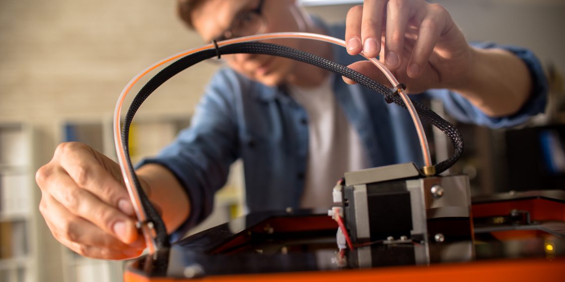 Close up of young man wearing glasses connecting cables to 3D printing machine while working on creative project in design studio