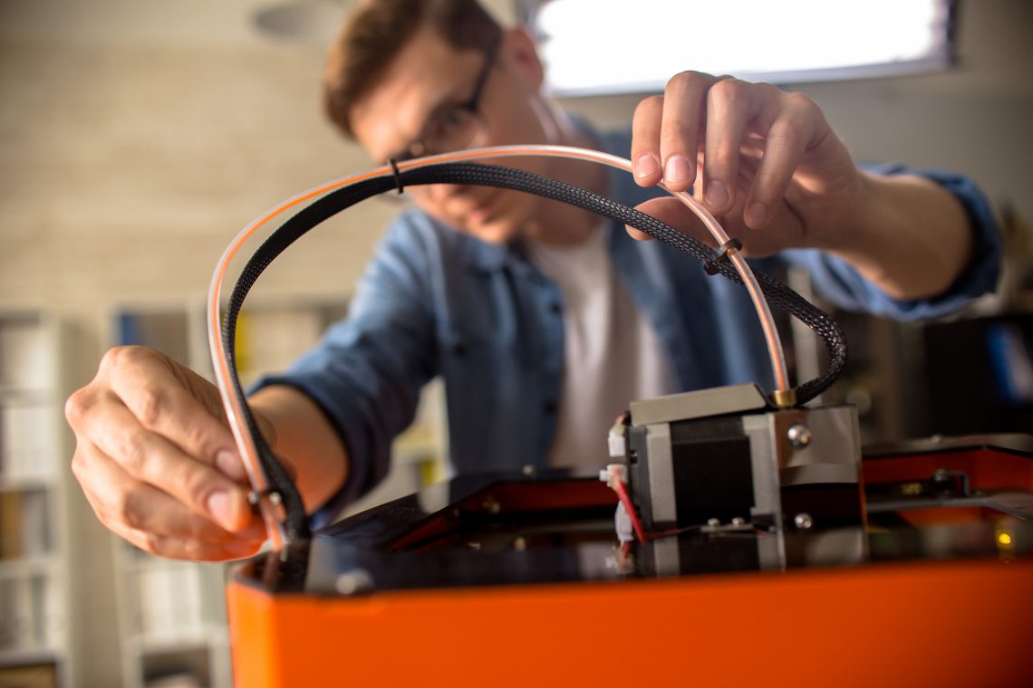 Close up of young man wearing glasses connecting cables to 3D printing machine while working on creative project in design studio