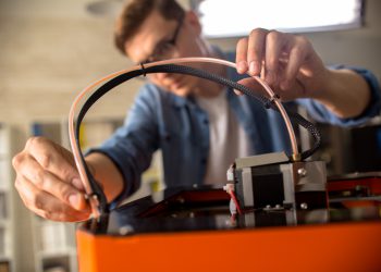 Close up of young man wearing glasses connecting cables to 3D printing machine while working on creative project in design studio