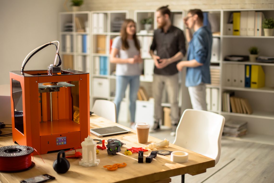 Portrait of three creative young people working in modern design studio, focus on 3D printer on table in foreground, copy space