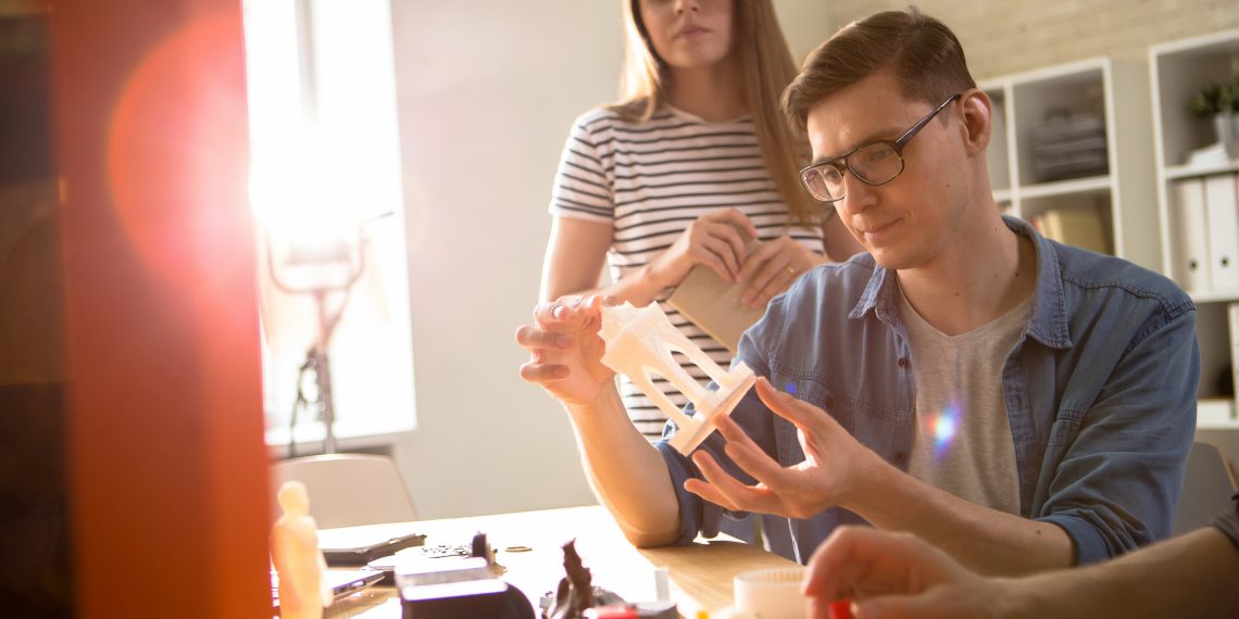 Confident young technician in eyeglasses carrying out quality inspection of 3D printed detail while having working meeting with colleagues at modern lab