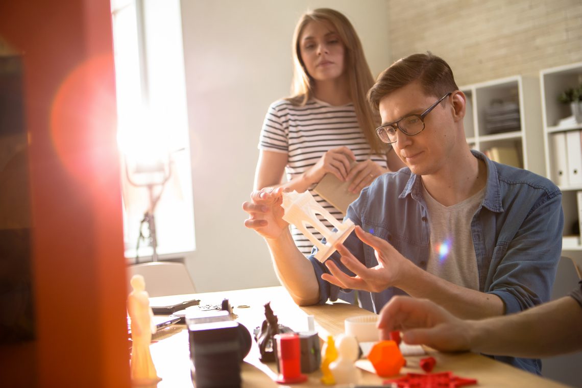 Confident young technician in eyeglasses carrying out quality inspection of 3D printed detail while having working meeting with colleagues at modern lab