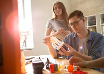 Confident young technician in eyeglasses carrying out quality inspection of 3D printed detail while having working meeting with colleagues at modern lab